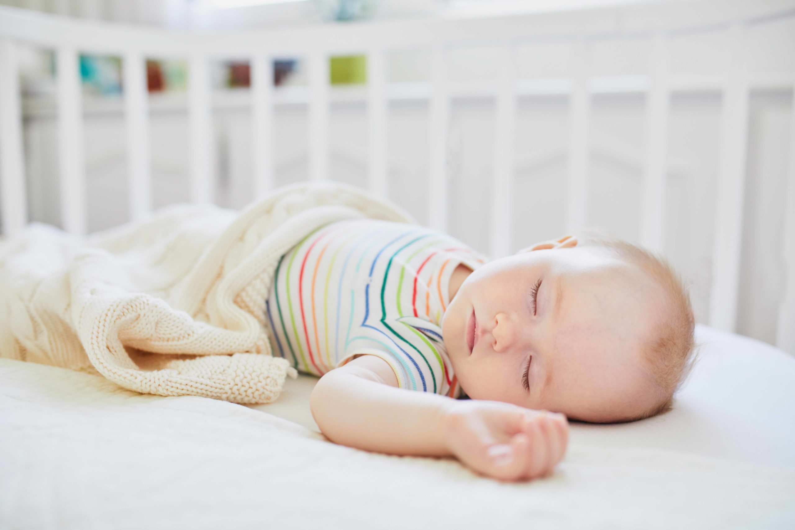 Adorable baby girl sleeping in co-sleeper crib attached to parents' bed. Little child having a day nap in cot. Sleep training concept. Infant kid in sunny nursery